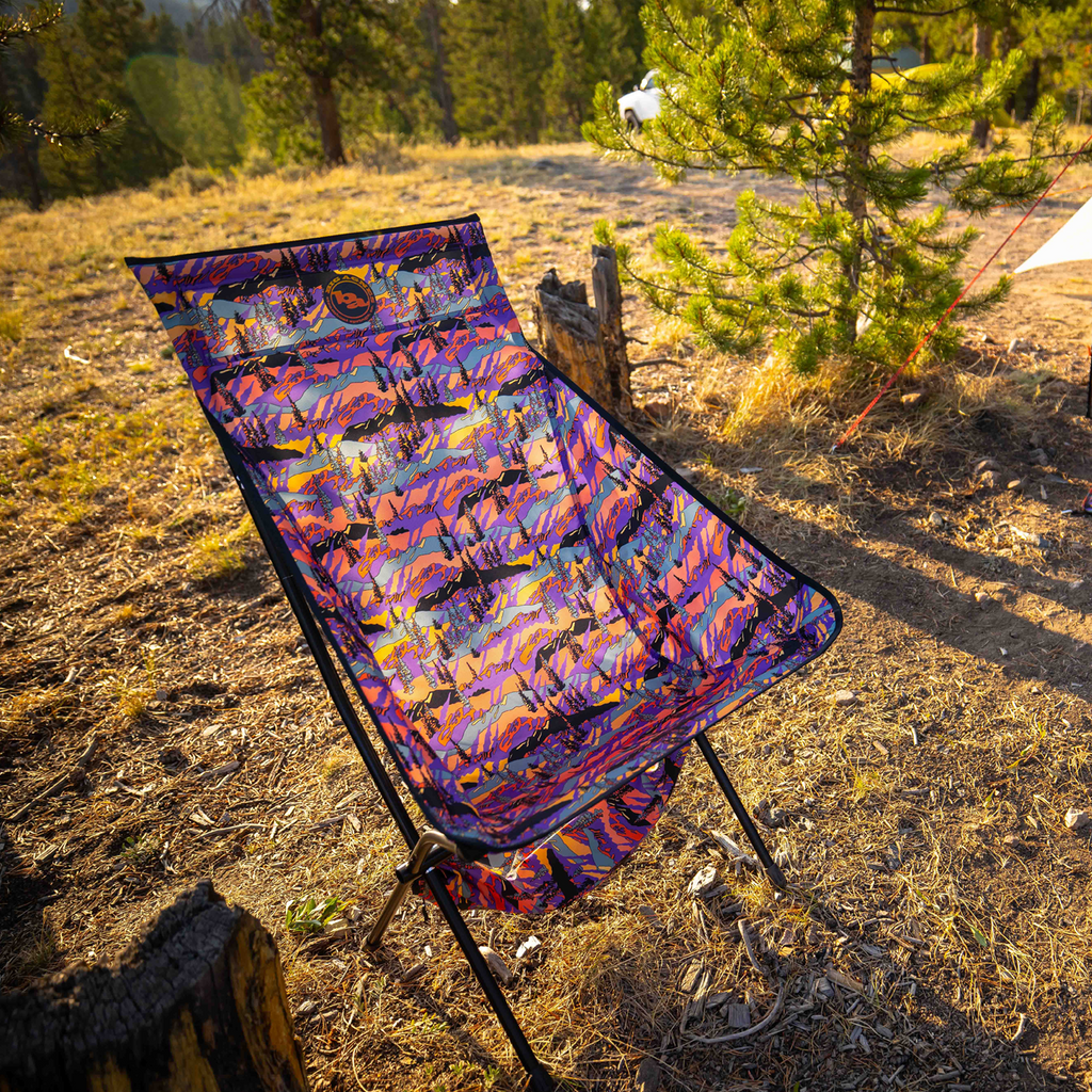 The Big Agnes Returns Big Six Camp Chair - Alpenglow, featuring a colorful, patterned design, is set up on dry grass near pine trees. Sunlight casts warm shadows on the chair and ground, creating an inviting outdoor atmosphere.