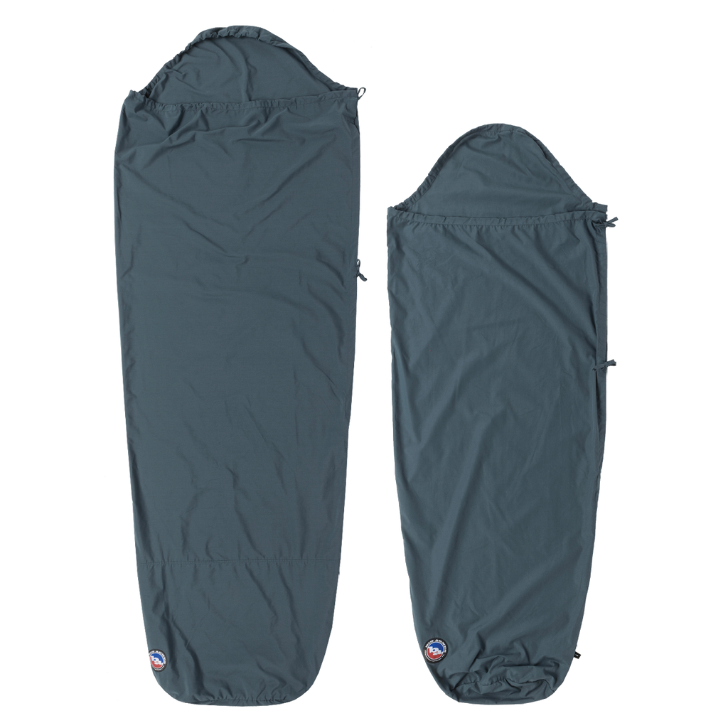 Two dark blue Big Agnes Cotton Sleeping Bag Liners are laid flat. The left is rectangular with a hood; the right is a tapered mummy style. Both feature a small logo at the bottom and are ideal for travel.