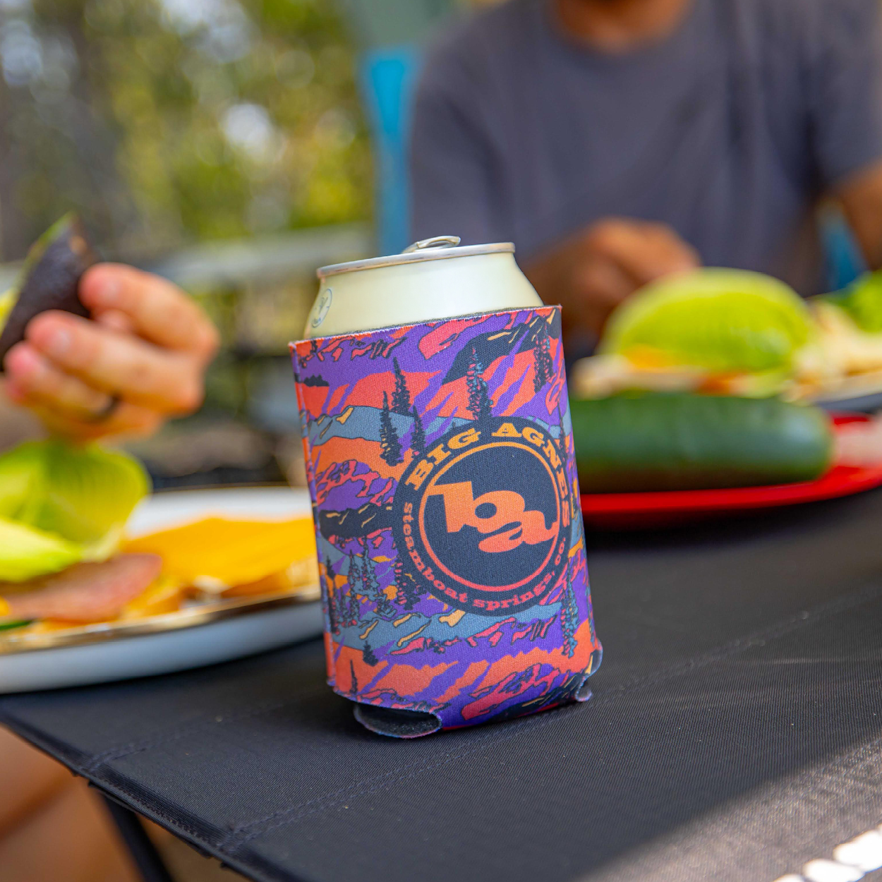 A close-up of a soda can in the limited edition Big Agnes Returns Alpenglow Koozie sits on a picnic table, with plates of food and two people blurred in the background.