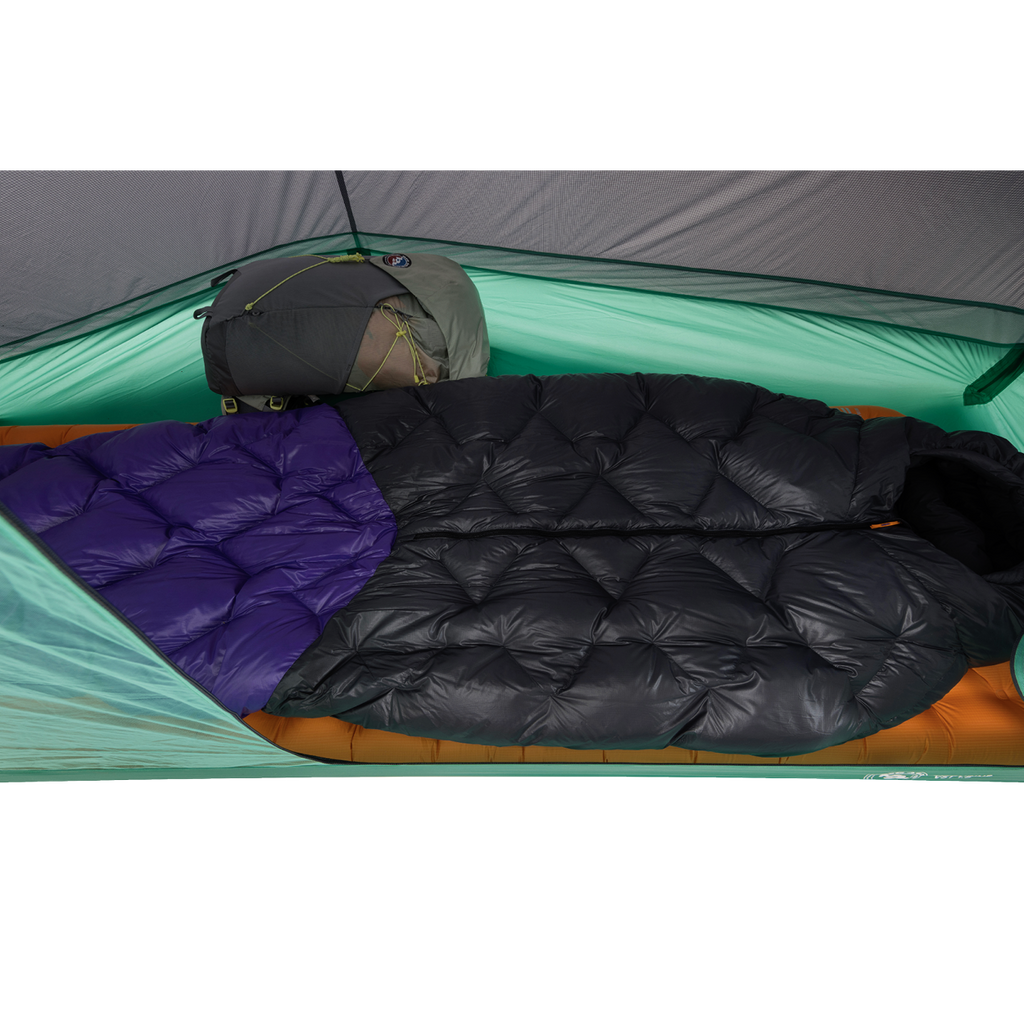 Two sleeping bags—one all black and one with a purple section—are side by side inside a green Big Agnes Pitchpine VST 1.5 tent, set up with an orange sleeping pad beneath and a gray backpack at the head for an ultralight shelter setup.