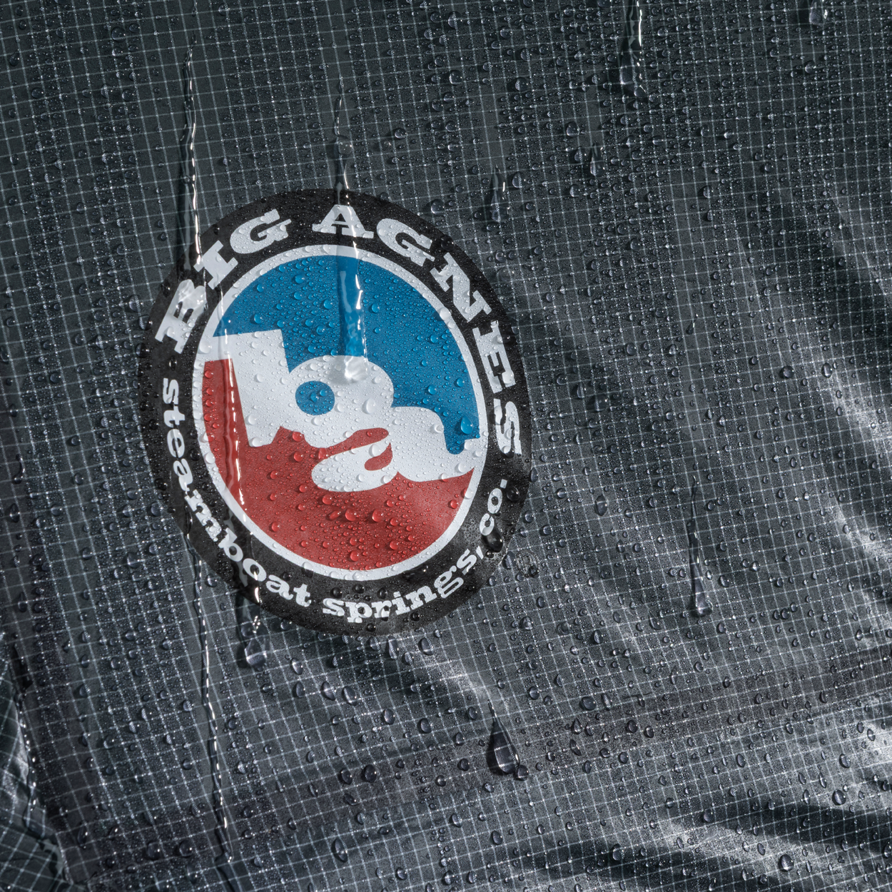 Close-up of wet, grid-patterned Hyperbead fabric with blue and red Big Agnes Returns logo, covered in droplets—ideal for use in the Pitchpine VST 1.5 tent and other ultralight shelters.