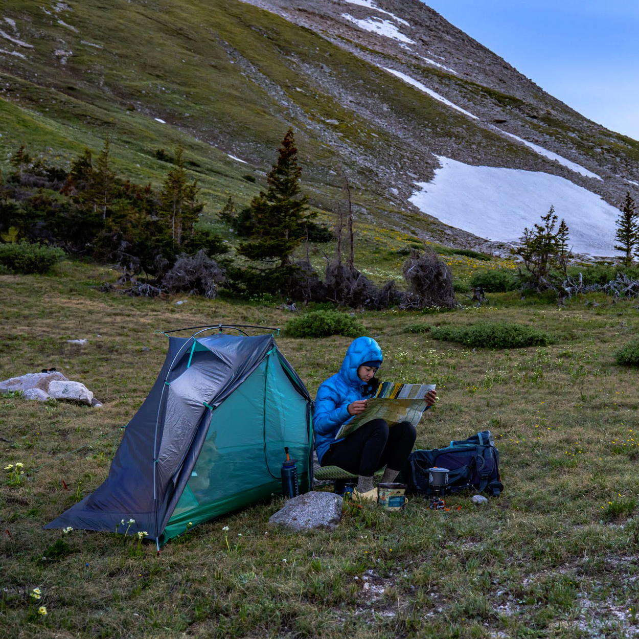 A person in a blue jacket sits beside a green Big Agnes Returns Pitchpine VST 1.5 tent, reading a map on a grassy mountain slope dotted with trees and snow. Ultralight camping gear made with Hyperbead fabric is nearby.