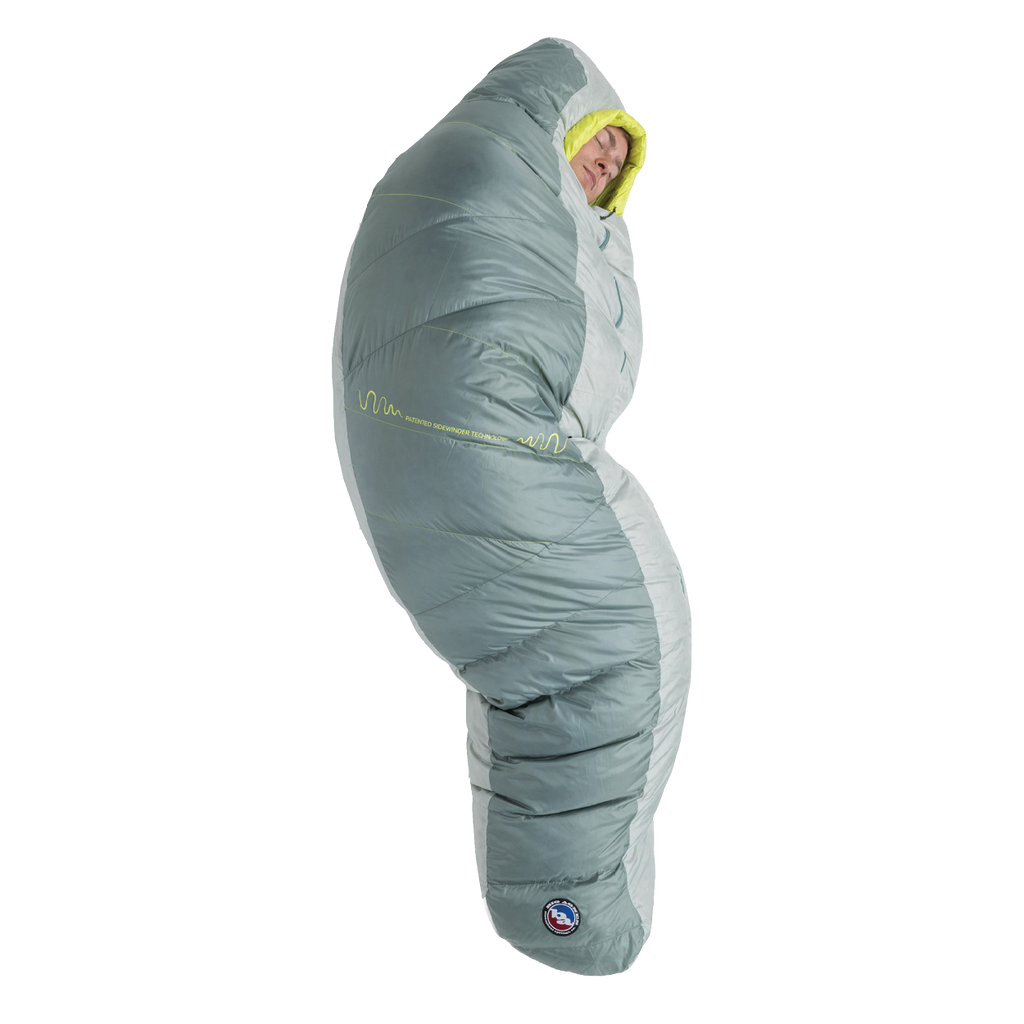 A person stands upright, fully zipped inside the Big Agnes Sidewinder 0˚ sleeping bag in gray and light green, featuring body-mapped insulation with only their face visible.