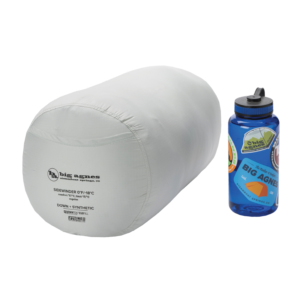 A light gray Big Agnes Sidewinder 0˚ sleeping bag with body-mapped insulation, packed in a cylindrical stuff sack, sits next to a blue sticker-covered Nalgene bottle on a white background.