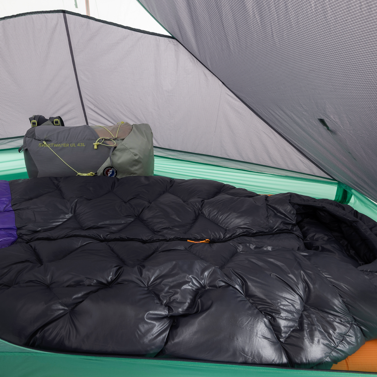 A cozy black sleeping bag lies inside a green Big Agnes Returns String Ridge VST 1.5 tent, with a gray backpack and gear by the back wall. Soft light filters through the mesh ceiling and walls of this ultralight shelter.