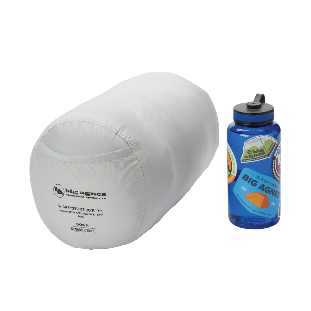 A packed Big Agnes Returns Women's Greystone 30° sleeping bag is shown next to a blue Nalgene water bottle for size comparison, both placed on a plain white background.
