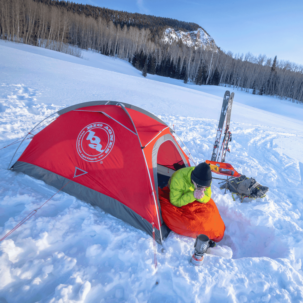 A person sits in a bright orange sleeping bag beside the red Big Agnes Shield 2 tent on snow, sipping from a mug. Skis, poles, and gear are nearby with bare trees, a snowy landscape, and a mountain in the background.