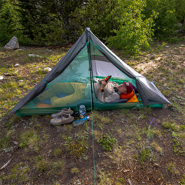 Inside a Big Agnes String Ridge VST 1.5 tent, a person relaxes on a sleeping pad with a book. Nearby, hiking shoes and a water bottle sit outside the tent on uneven grass, surrounded by green trees and bushes.